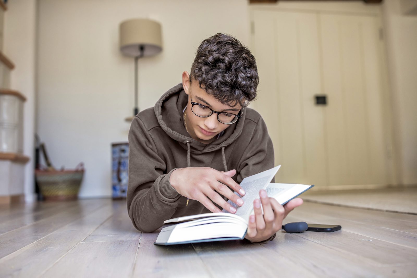 A teenager in spectacles reading a book on the floor at home with a phone beside them