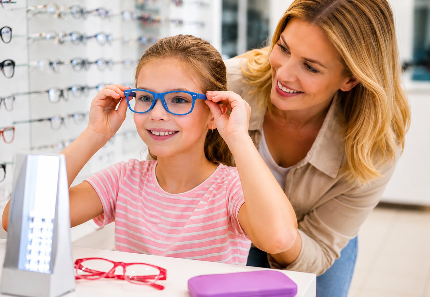 A girl trying on a pair of blue spectacle frames while an optician helps her, in an optical shop