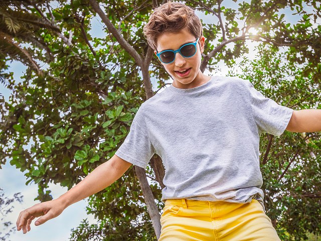A boy outdoors under sunlit trees, the lenses of his blue-framed spectacles darkened in the bright light