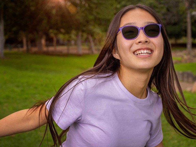 A girl laughing in a sunlit park, wearing spectacles with dark purple-framed tinted lenses